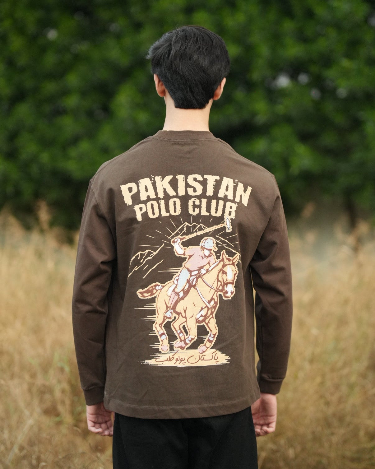 Person wearing a brown 'Pakistan Polo Club' long-sleeve shirt with a graphic design outdoors.